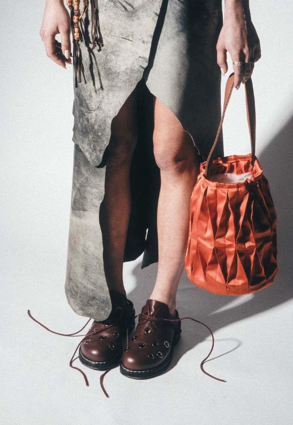 Folded rust bucket bag carried beside a raw-edged skirt and brown leather shoes